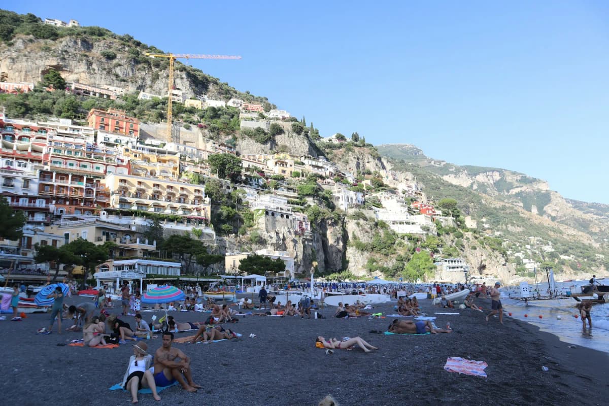 Sunbathers on a dark-sand beach in Positano below hotels and houses on the cliff
