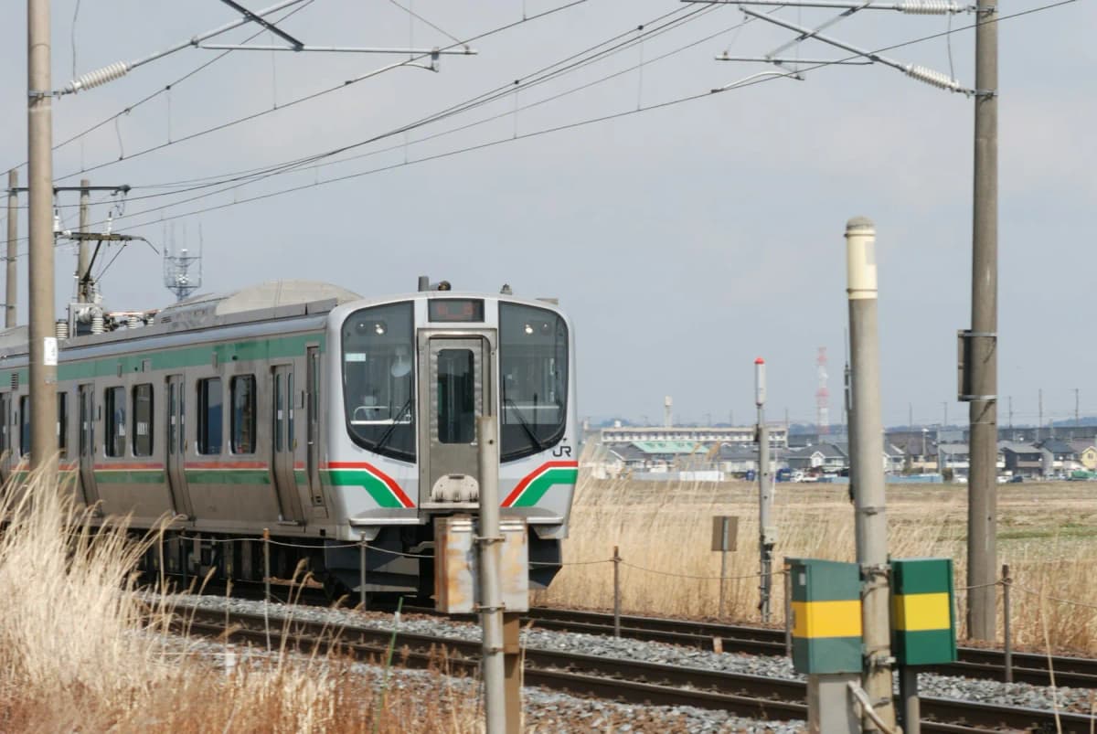 A local train running through flat countryside in Japan under overhead power lines