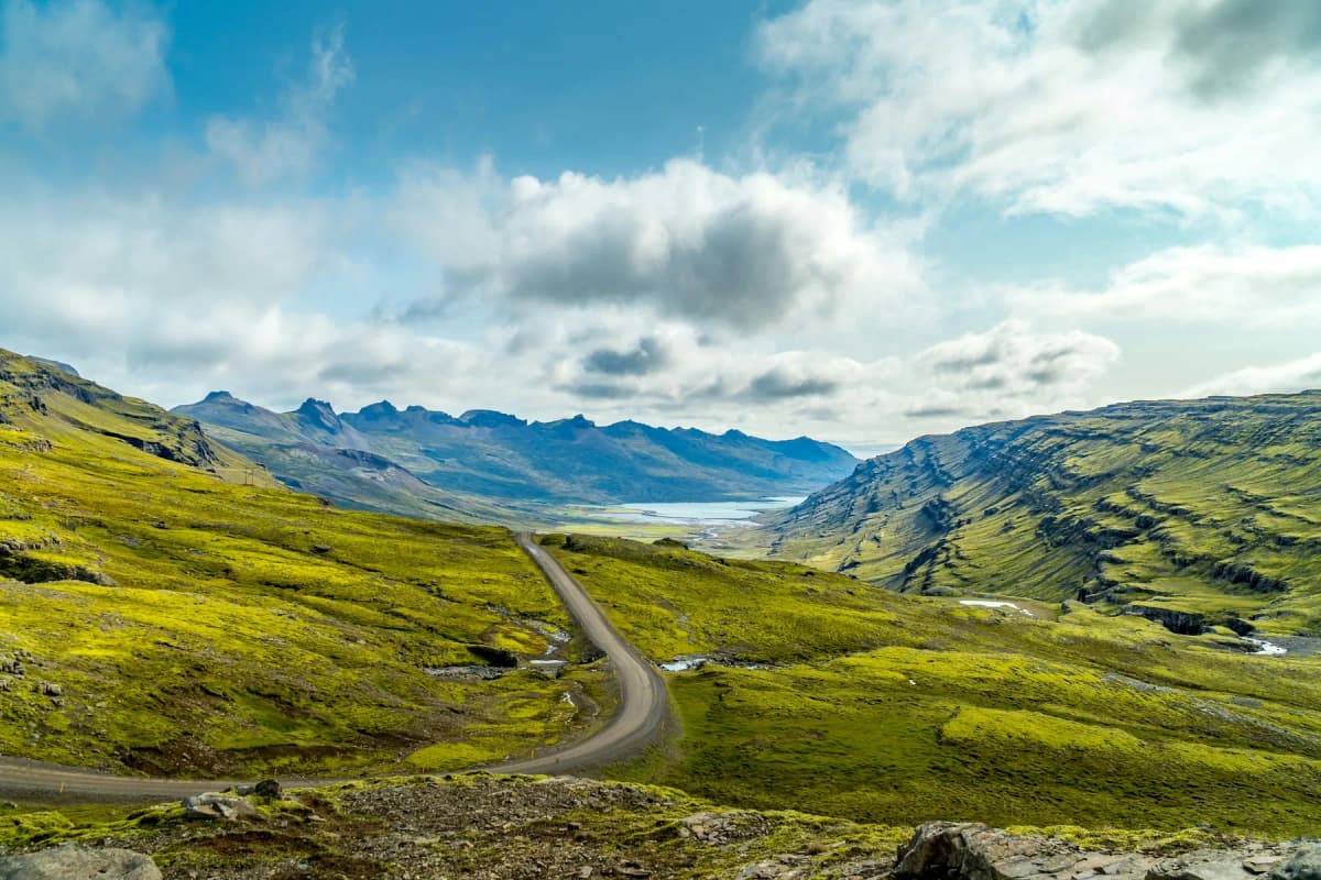 A road winding down a green valley towards a fjord with mountains in the distance