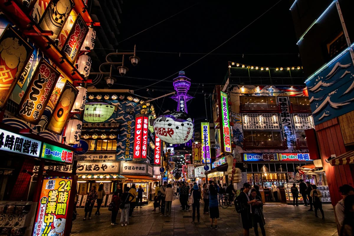 Lanterns and neon signs on a street in Osaka's Shinsekai district with Tsutenkaku Tower behind
