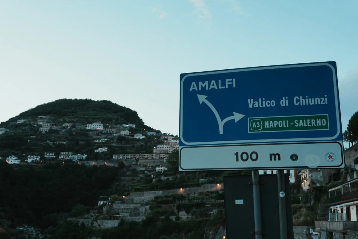 A blue Italian road sign pointing to Amalfi, with hills in the background