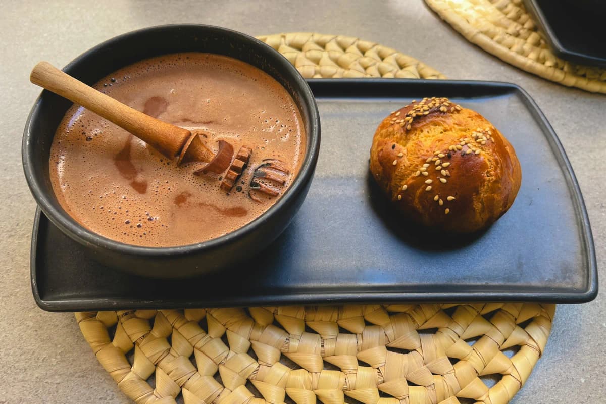 A bowl of frothy Oaxacan hot chocolate with a wooden stirrer and a round sweet bread