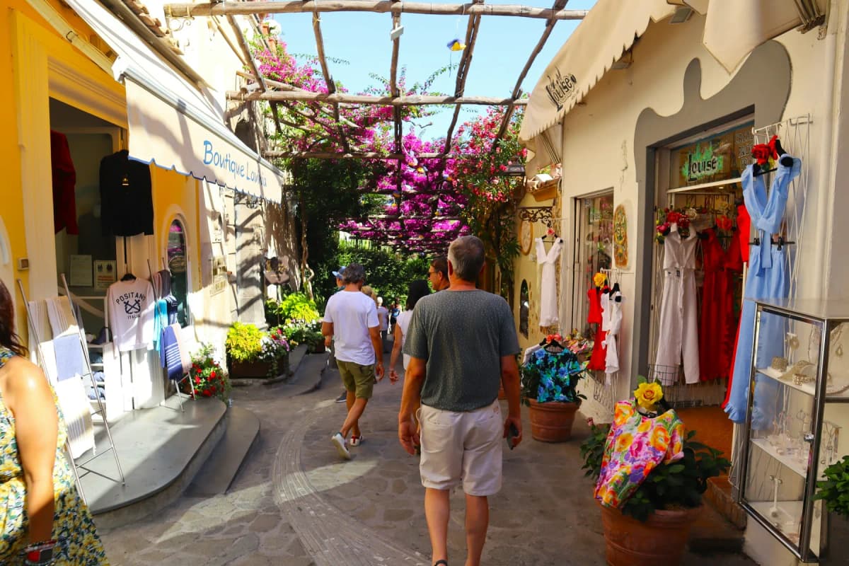 Shoppers walking along a lane of clothing boutiques under a bougainvillea pergola in Positano