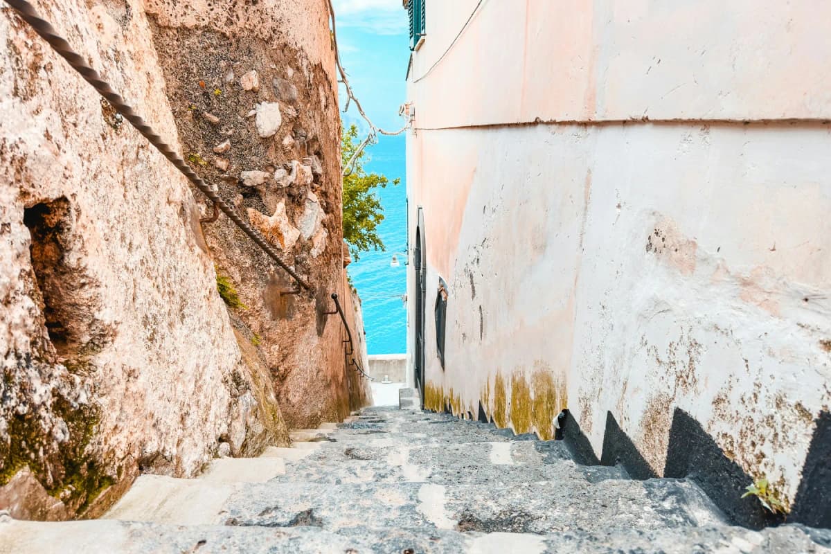 A narrow stone stairway between old walls in Positano with a glimpse of blue sea at the end