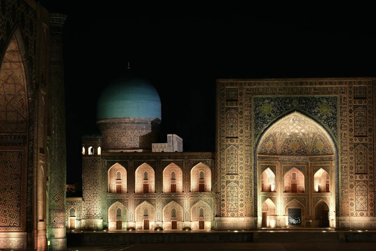 The Registan's madrasahs lit up at night in Samarkand, with a turquoise dome glowing above the arches